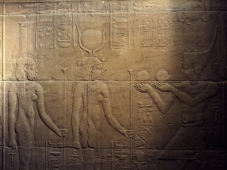 Ancient Egyptian stone relief depicting three figures in traditional attire. The central figure wears a headdress with a sun disk and horns, symbolizing divinity. Hieroglyphics accompany the scene.