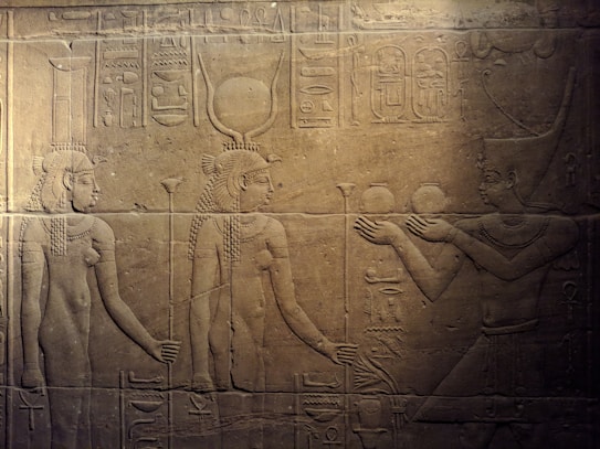 Ancient Egyptian stone relief depicting three figures in traditional attire. The central figure wears a headdress with a sun disk and horns, symbolizing divinity. Hieroglyphics accompany the scene.