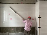 A worker wearing a red hard hat and a pink shirt is measuring tiles on a wall with a long tool. The tiled wall has a light, textured pattern, and there are pink accent squares positioned at intervals. The unfinished lower section reveals some concrete.
