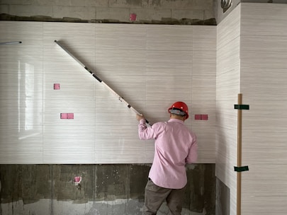A worker wearing a red hard hat and a pink shirt is measuring tiles on a wall with a long tool. The tiled wall has a light, textured pattern, and there are pink accent squares positioned at intervals. The unfinished lower section reveals some concrete.