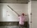A worker wearing a red hard hat and a pink shirt is measuring tiles on a wall with a long tool. The tiled wall has a light, textured pattern, and there are pink accent squares positioned at intervals. The unfinished lower section reveals some concrete.