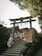 A group of people wearing traditional Japanese attire are ascending stone steps leading to a Shinto shrine gate, surrounded by lush greenery. The torii gate is prominently displayed, with a wooden plaque hanging in the center and wooden décor underneath. Signs with Japanese characters are visible alongside the steps.