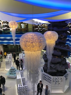 The image depicts the interior of a modern, well-lit space with a marine theme, featuring large, luminous jellyfish-like decorations hanging from the ceiling. Beneath them, several people are walking around and observing various items displayed on white shelves and stands. The background includes a large artificial rock structure and wavy blue-and-white decorative elements.