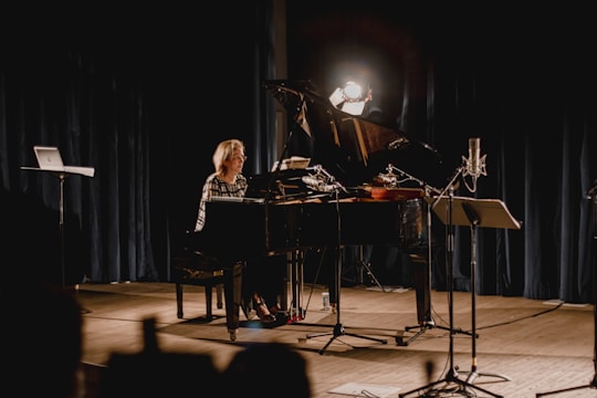 A musician seated at a grand piano on a dimly lit stage, surrounded by microphones and a music stand. Stage lights create a focused spotlight on the pianist, with dark curtains as a backdrop adding to the intimate concert setting.