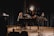 A musician seated at a grand piano on a dimly lit stage, surrounded by microphones and a music stand. Stage lights create a focused spotlight on the pianist, with dark curtains as a backdrop adding to the intimate concert setting.