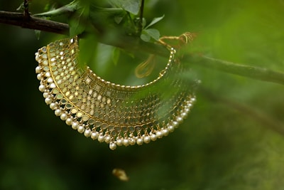 A traditional gold choker necklace adorned with intricate patterns and small pearls, laid gently on a tree branch surrounded by lush green leaves.