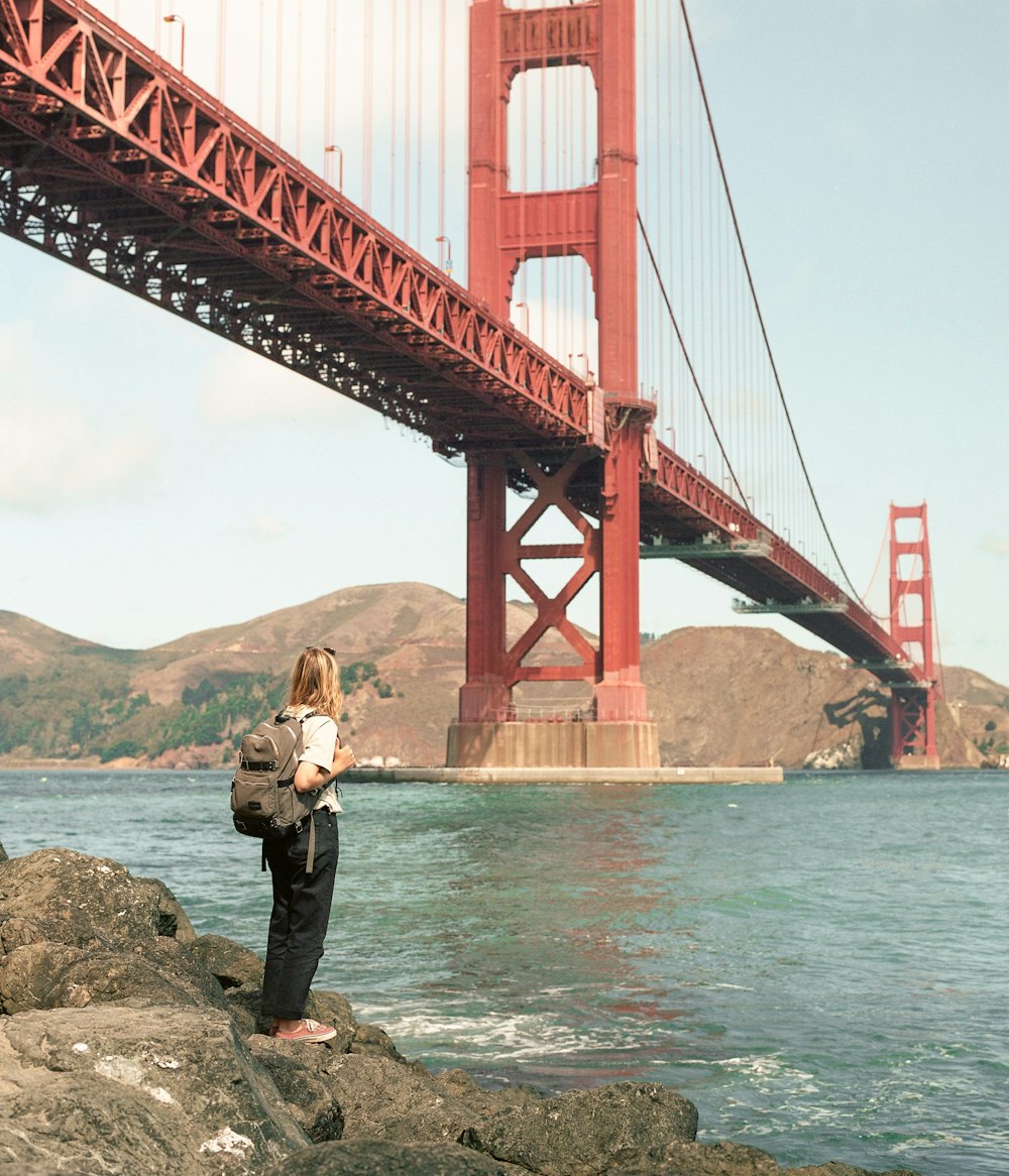 uma pessoa de pé sobre rochas perto da água e uma ponte