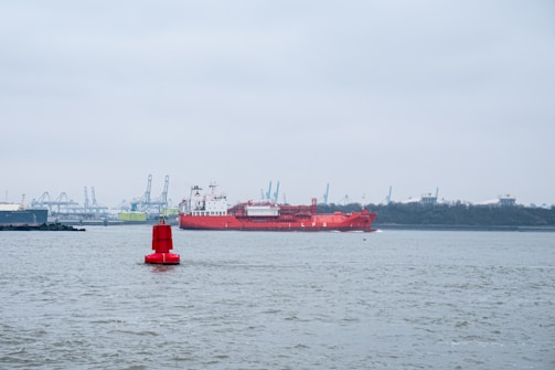 A large red LNG ship sails across a calm body of water, with a bright red buoy in the foreground. The horizon features industrial cranes, suggesting a port or harbor setting, under a cloudy gray sky.