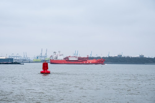 A large red LNG ship sails across a calm body of water, with a bright red buoy in the foreground. The horizon features industrial cranes, suggesting a port or harbor setting, under a cloudy gray sky.