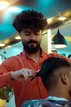 A hairstylist with curly hair and a beard is attentively cutting a client's hair in a modern barbershop. The barber is wearing an orange shirt and is working with a comb and scissors. The salon is well-lit with contemporary lighting fixtures visible in the background.