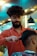 A hairstylist with curly hair and a beard is attentively cutting a client's hair in a modern barbershop. The barber is wearing an orange shirt and is working with a comb and scissors. The salon is well-lit with contemporary lighting fixtures visible in the background.