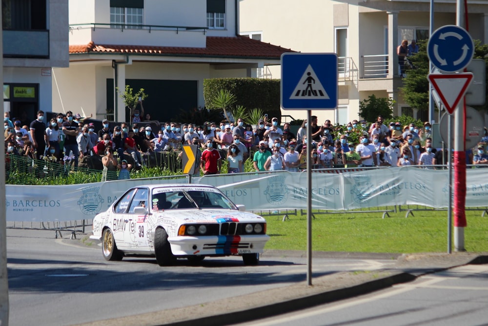 a white car driving down a street past a crowd of people