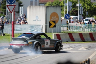 A vintage sports car is drifting on a road with smoke coming from the tires, while spectators watch from behind barriers. The environment is set in an urban area during a car rally event.