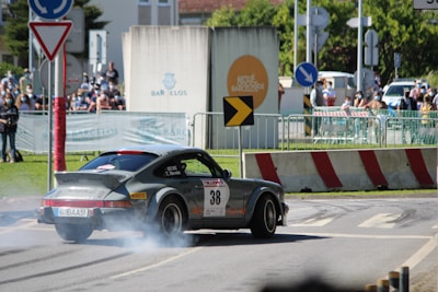 A vintage sports car is drifting on a road with smoke coming from the tires, while spectators watch from behind barriers. The environment is set in an urban area during a car rally event.