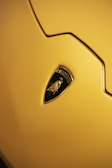 A close-up of the emblem from a Lamborghini car on a sleek yellow surface. The emblem features a black and gold design with a shield shape, showcasing the signature Lamborghini bull.