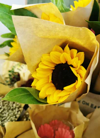 A rustic bouquet featuring wildflowers and vibrant sunflowers, wrapped in brown craft paper.