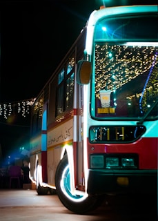 A brightly lit bus is parked during the night, decorated with festive lights that create a colorful and lively ambiance. Inside the bus, there are visible lights and decorations, and there is a sign on the front windshield promoting a beverage that is 'Available Now'. The bus has a red, white, and blue color scheme and the pavement it is parked on is visible, with some faintly lit objects near the bus.