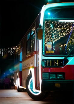 A brightly lit bus is parked during the night, decorated with festive lights that create a colorful and lively ambiance. Inside the bus, there are visible lights and decorations, and there is a sign on the front windshield promoting a beverage that is 'Available Now'. The bus has a red, white, and blue color scheme and the pavement it is parked on is visible, with some faintly lit objects near the bus.