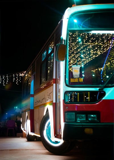 A brightly lit bus is parked during the night, decorated with festive lights that create a colorful and lively ambiance. Inside the bus, there are visible lights and decorations, and there is a sign on the front windshield promoting a beverage that is 'Available Now'. The bus has a red, white, and blue color scheme and the pavement it is parked on is visible, with some faintly lit objects near the bus.