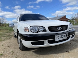 A white sedan car is parked on a gravel surface under a partly cloudy sky. The vehicle has a sleek design with prominent headlights. Its front license plate reads UG-43 88, with Chile indicated as the location.