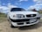 A white sedan car is parked on a gravel surface under a partly cloudy sky. The vehicle has a sleek design with prominent headlights. Its front license plate reads UG-43 88, with Chile indicated as the location.