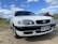 A white sedan car is parked on a gravel surface under a partly cloudy sky. The vehicle has a sleek design with prominent headlights. Its front license plate reads UG-43 88, with Chile indicated as the location.