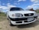 A white sedan car is parked on a gravel surface under a partly cloudy sky. The vehicle has a sleek design with prominent headlights. Its front license plate reads UG-43 88, with Chile indicated as the location.