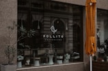 A storefront with a large glass window displaying the logo of Pollito Cheesecake. There are potted plants and a white stool visible within the window display. An orange umbrella is partially seen on the right side of the image, and the background includes reflections of an ornate building facade. A man is also partially visible in a black and white poster on the right.