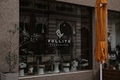 A storefront with a large glass window displaying the logo of Pollito Cheesecake. There are potted plants and a white stool visible within the window display. An orange umbrella is partially seen on the right side of the image, and the background includes reflections of an ornate building facade. A man is also partially visible in a black and white poster on the right.