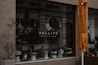 A storefront with a large glass window displaying the logo of Pollito Cheesecake. There are potted plants and a white stool visible within the window display. An orange umbrella is partially seen on the right side of the image, and the background includes reflections of an ornate building facade. A man is also partially visible in a black and white poster on the right.