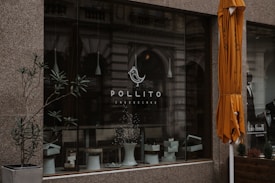 A storefront with a large glass window displaying the logo of Pollito Cheesecake. There are potted plants and a white stool visible within the window display. An orange umbrella is partially seen on the right side of the image, and the background includes reflections of an ornate building facade. A man is also partially visible in a black and white poster on the right.