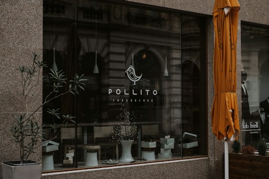A storefront with a large glass window displaying the logo of Pollito Cheesecake. There are potted plants and a white stool visible within the window display. An orange umbrella is partially seen on the right side of the image, and the background includes reflections of an ornate building facade. A man is also partially visible in a black and white poster on the right.