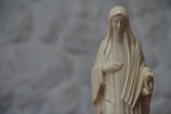 A beige statue of a woman with a serene expression, wearing a long robe and veil, with one hand on her chest and the other slightly extended. The background is a soft, out-of-focus gray pattern.