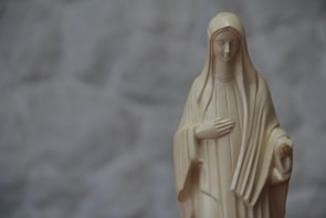 A beige statue of a woman with a serene expression, wearing a long robe and veil, with one hand on her chest and the other slightly extended. The background is a soft, out-of-focus gray pattern.