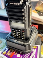 A handheld radio transceiver, labeled 'BAOFENG', is positioned on a charging dock. The radio features multiple buttons, a small display screen, and is set against a backdrop with an isopropyl alcohol wipe packet partially visible.