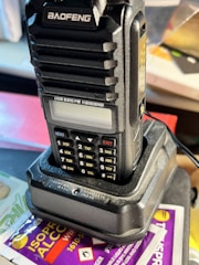 A handheld radio transceiver, labeled 'BAOFENG', is positioned on a charging dock. The radio features multiple buttons, a small display screen, and is set against a backdrop with an isopropyl alcohol wipe packet partially visible.