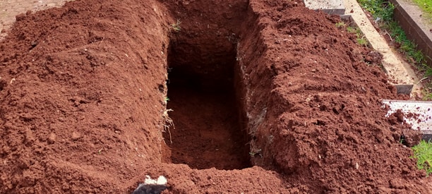 A freshly dug rectangular hole in the ground with mounds of dark brown soil surrounding it. The earth appears recently disturbed, and some grass and paving stones are visible on the right side.