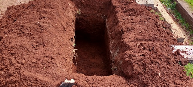 A freshly dug rectangular hole in the ground with mounds of dark brown soil surrounding it. The earth appears recently disturbed, and some grass and paving stones are visible on the right side.