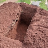 A freshly dug rectangular hole in the ground with brown, loose soil piled around its edges. Nearby, the corner of a gravestone is visible on a patch of green grass.