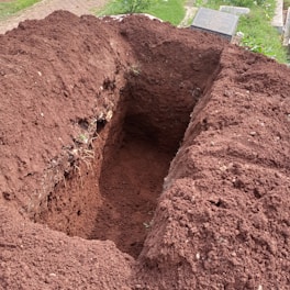 A freshly dug rectangular hole in the ground with brown, loose soil piled around its edges. Nearby, the corner of a gravestone is visible on a patch of green grass.