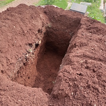 A freshly dug rectangular hole in the ground with brown, loose soil piled around its edges. Nearby, the corner of a gravestone is visible on a patch of green grass.