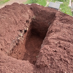 A freshly dug rectangular hole in the ground with brown, loose soil piled around its edges. Nearby, the corner of a gravestone is visible on a patch of green grass.