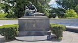 A bronze statue depicting two men, with one cradling the other, is mounted on a large, dark stone base. The base has the inscriptions 'FRIEND TO FRIEND' and 'A Brotherhood Undivided' along with a Masonic symbol. The monument is surrounded by lush greenery and a circular stone wall engraved with state names.