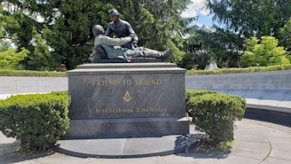 A bronze statue depicting two men, with one cradling the other, is mounted on a large, dark stone base. The base has the inscriptions 'FRIEND TO FRIEND' and 'A Brotherhood Undivided' along with a Masonic symbol. The monument is surrounded by lush greenery and a circular stone wall engraved with state names.