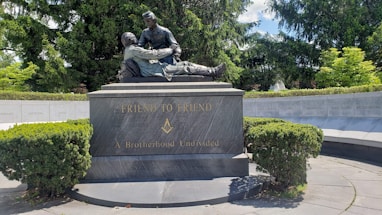 A bronze statue depicting two men, with one cradling the other, is mounted on a large, dark stone base. The base has the inscriptions 'FRIEND TO FRIEND' and 'A Brotherhood Undivided' along with a Masonic symbol. The monument is surrounded by lush greenery and a circular stone wall engraved with state names.