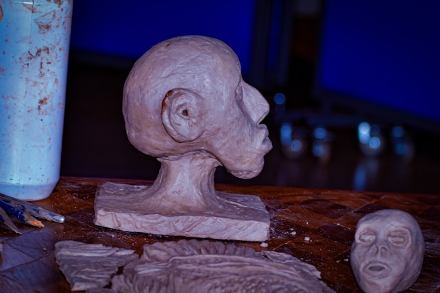 A close-up view of clay sculptures on a table, featuring a prominently molded head in profile with exaggerated facial features on a pedestal, accompanied by a smaller flat face sculpture and various clay tools. The background shows an indistinct blur, hinting at a studio or artist's workspace.