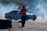 A motorsport event featuring a car drifting with smoke billowing from the tires. A person wearing an orange safety vest stands nearby taking photos, while red tents can be seen in the background.