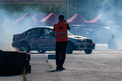 A motorsport event featuring a car drifting with smoke billowing from the tires. A person wearing an orange safety vest stands nearby taking photos, while red tents can be seen in the background.