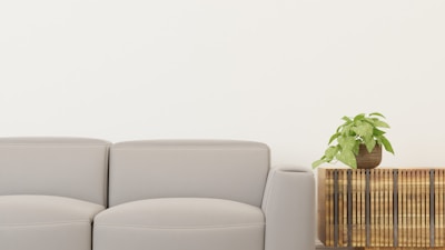 A modern, minimalist living space featuring a light gray sofa on the left and a wooden side table with vertical slats on the right. A green potted plant sits on top of the side table, adding a touch of nature to the scene. The backdrop is a plain, light-colored wall.