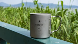A stainless steel mug with the brand name 'snow peak' inscribed on it sits on a green railing. In the background, there is lush green foliage and some distant mountains under a clear blue sky.